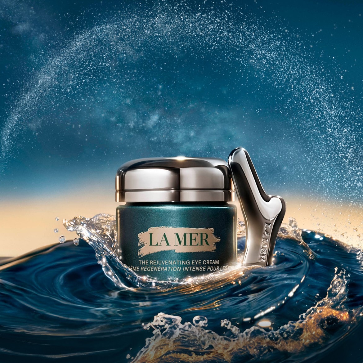 „La Mer The Rejuvenating Eye Cream“ žalsvai mėlyname stikliniame indelyje su sidabriniu dangteliu ir aplikatoriumi, apsuptas purslų.