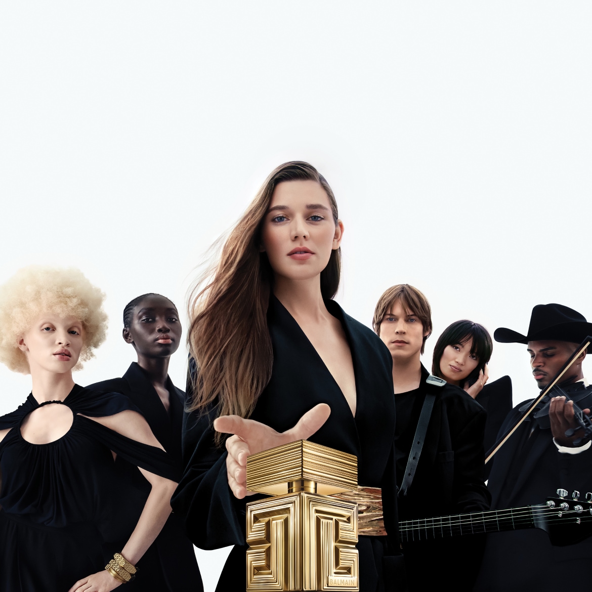 Moteris juodais drabužiais laiko auksinį „Balmain“ kvepalų buteliuką, o už jos stovi įvairūs žmonės, taip pat apsirengę juodai, įskaitant muzikantus su smuiku ir gitara.