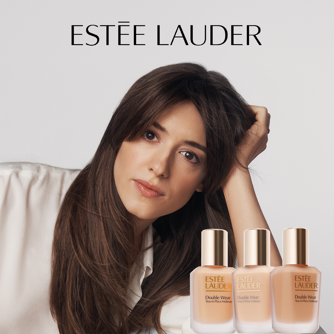 Šalia moters su šviesiu makiažu matomi trys „Estée Lauder Double Wear Stay-in-Place Makeup“ buteliukai su auksiniais dangteliais.