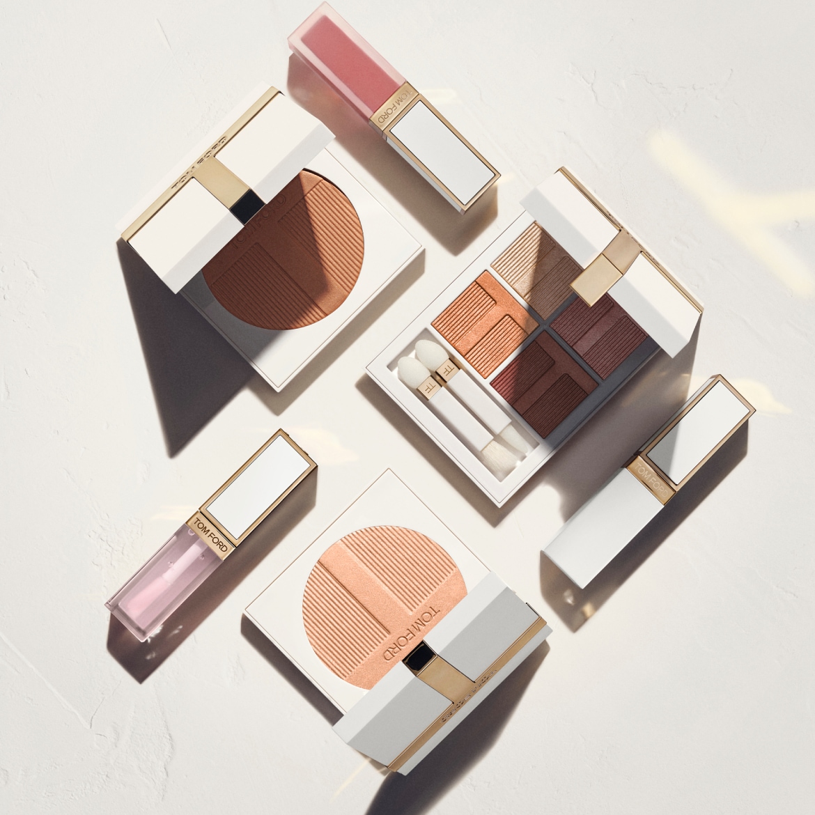Prabangiai išdėstyti „Tom Ford“ makiažo produktai – bronzantas, akių šešėlių paletė, lūpų blizgiai ir lūpdažiai – švelnių nude ir persikinių tonų, ant šviesaus neutralaus fono.