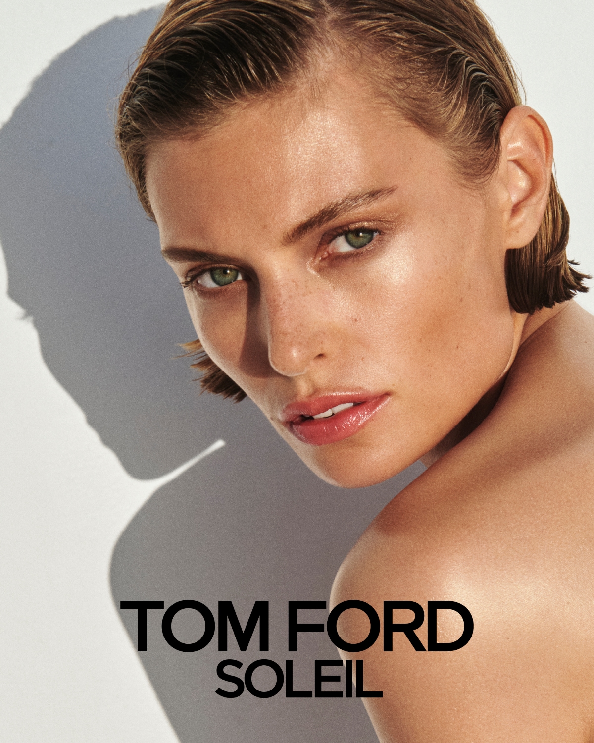 Asmuo su švytinčia oda ir atgal sušukuotais plaukais žiūri į kamerą, nuogas petys apšviestas saulės šviesa, šone matomas ryškus šešėlis. Apačioje tekstas „TOM FORD SOLEIL“.