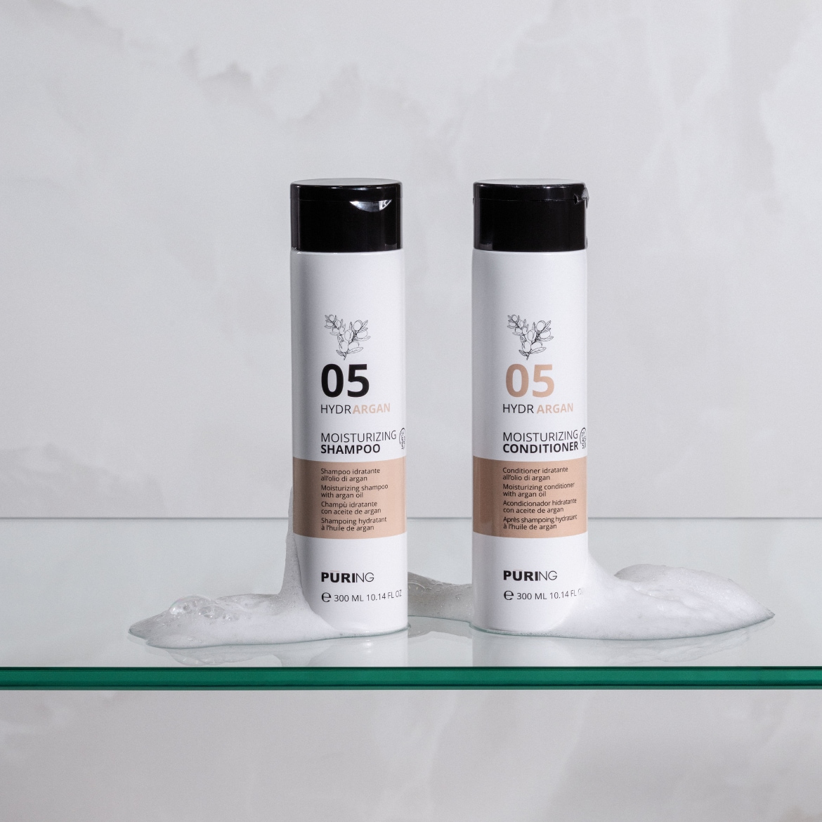 Du baltos „Puring HydrArgan“ šampūno ir kondicionieriaus buteliukai su juodais dangteliais, kurių kiekvieno talpa 300 ml, stovi ant stiklinės lentynos, o aplink juos matyti baltos putos.