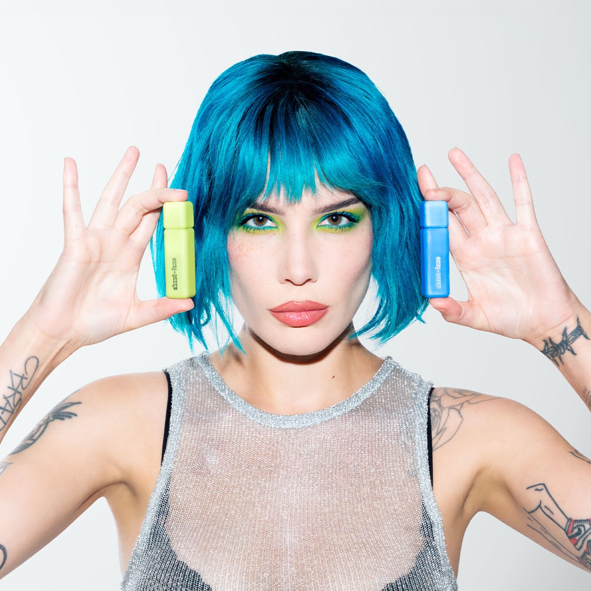 Moteris mėlynais plaukais laiko žalią ir mėlyną „about-face“ buteliukus, demonstruodama prekės ženklo spalvų pasirinkimą.