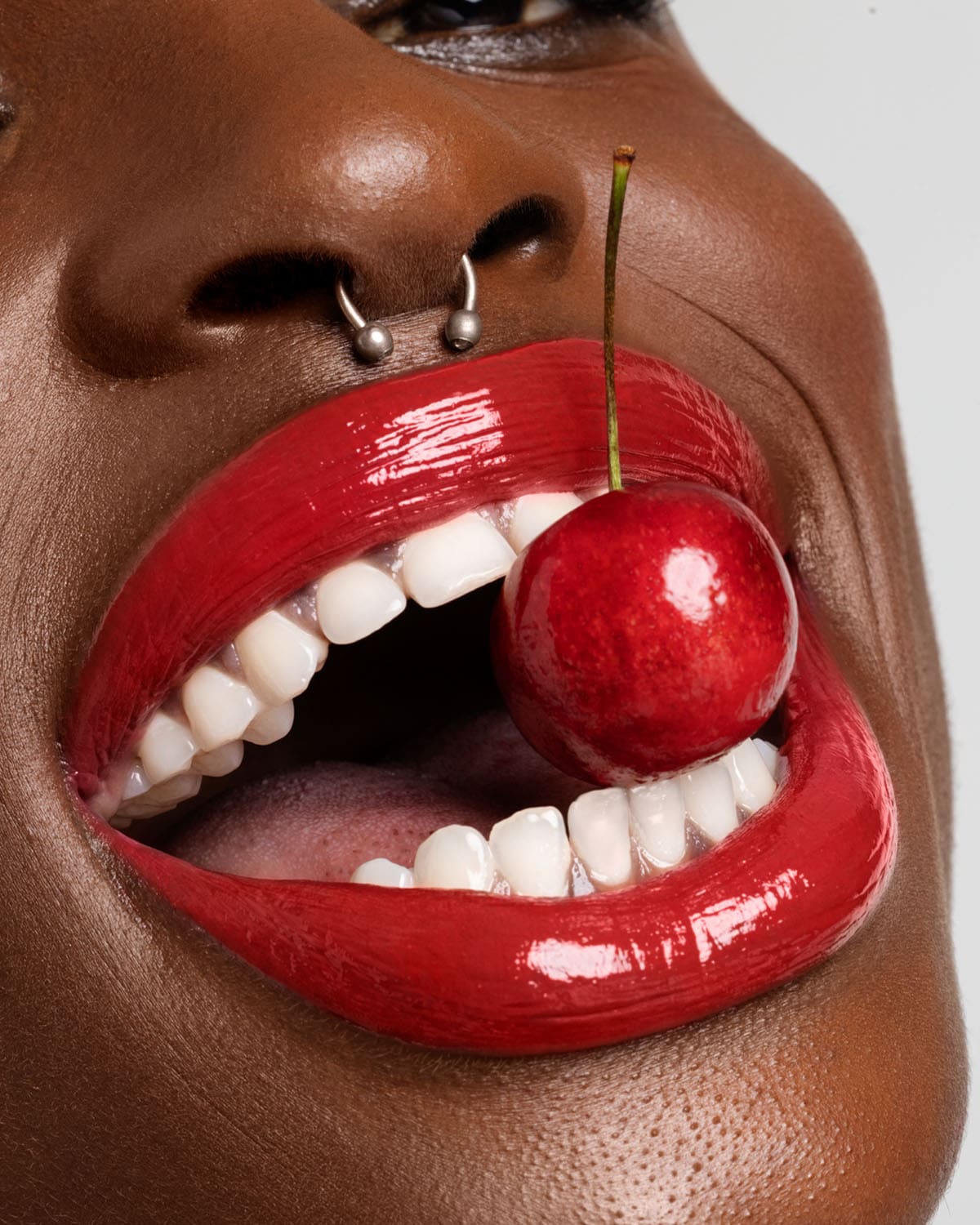 Tamsiaodė moteris su sidabriniu auskaru nosyje laiko vyšnią burnoje, kurios lūpos padažytos ryškiai raudonu blizgiu.