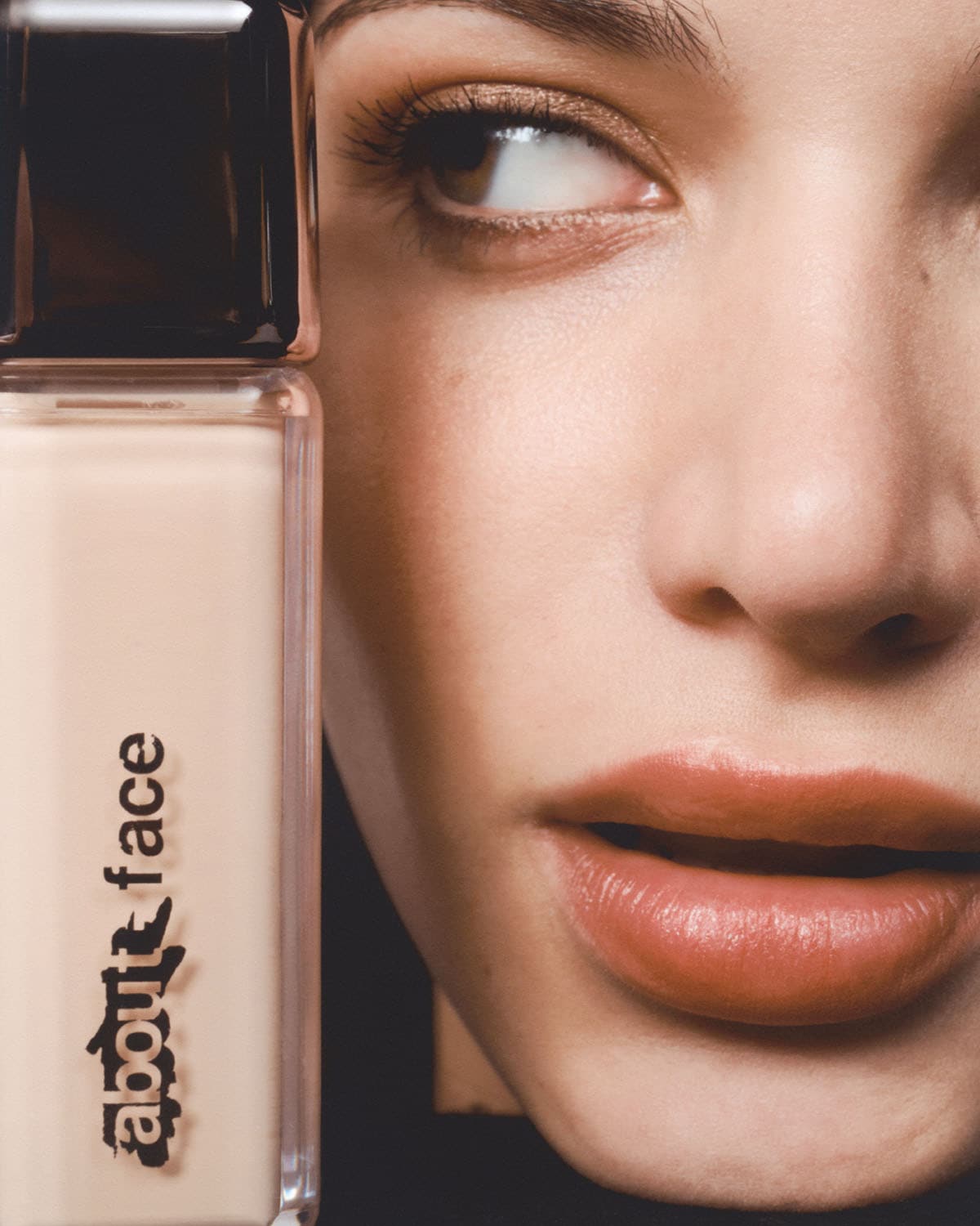 Šalia moters veido matomas „about-face“ pagrindo buteliukas su juodu dangteliu ir skaidriu korpusu, užpildytu smėlio spalvos skysčiu.