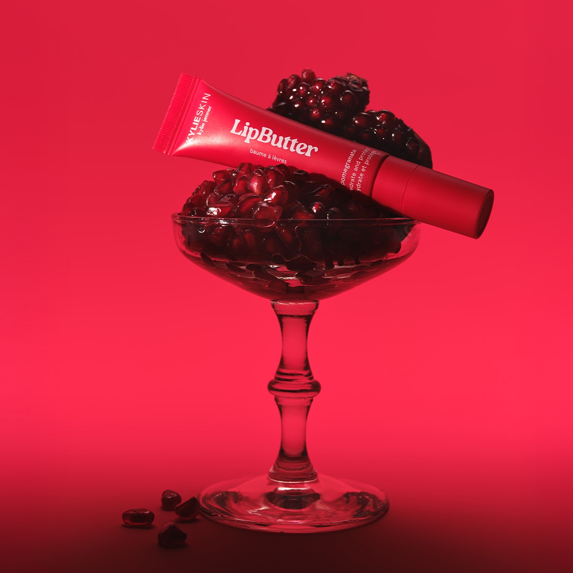 Raudonas „KylieSkin LipButter“ tūbelė guli ant taurės, pripildytos granatų sėklų, ant raudono fono.