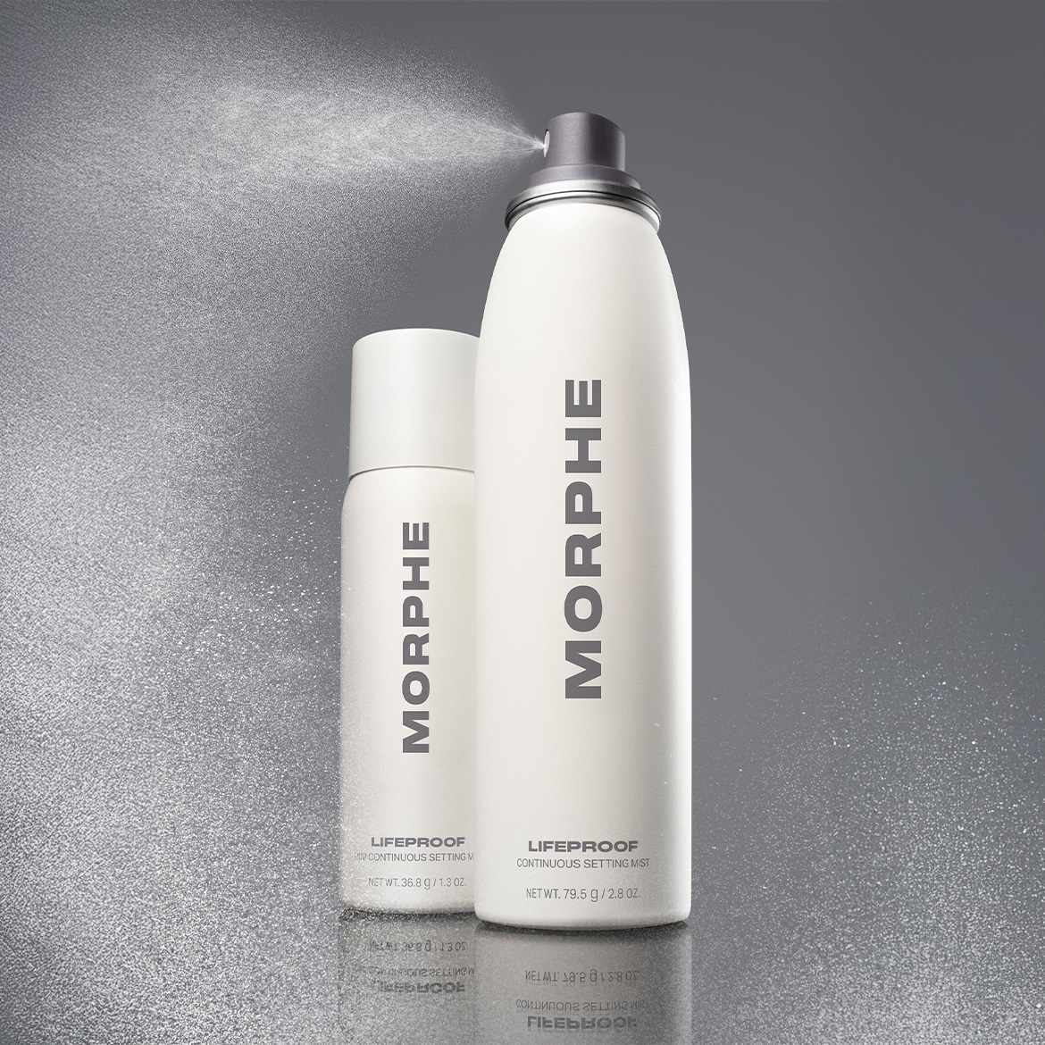 Du balto fono matosi du "Morphe" fiksavimo purškikliai, didesnis ir mažesnis, purškiami į orą, kad pademonstruotų savo smulkią dulksną.