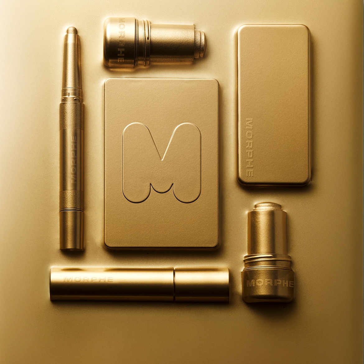 Auksinės spalvos „Morphe“ kosmetikos rinkinys, įskaitant akių pieštuką, lūpų dažus ir makiažo pagrindą, viskas išdėstyta ant auksinio paviršiaus.
