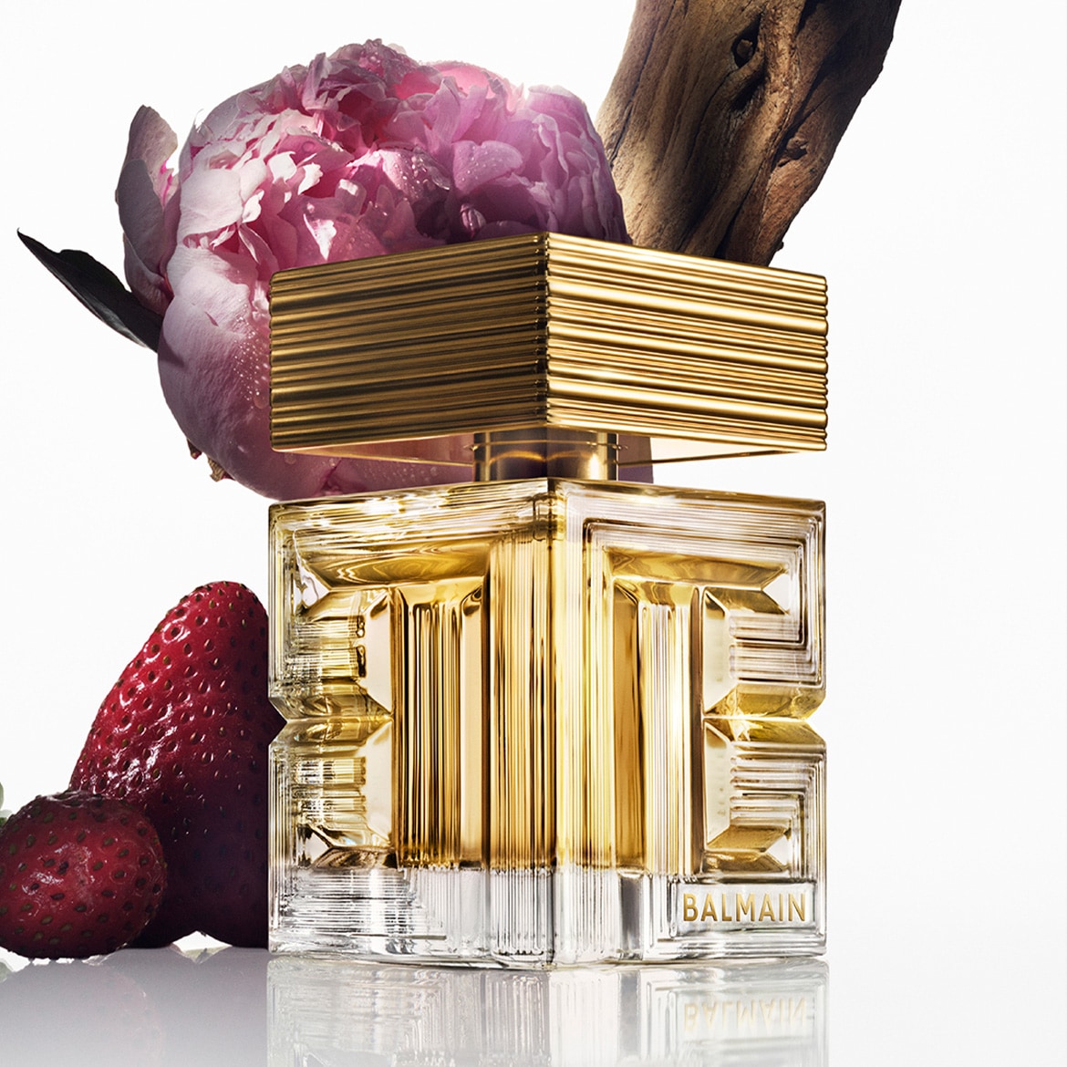 Auksinis Balmain kvepalų buteliukas su braškėmis ir bijūnu šalia, suteikiantis prabangos ir gamtos pojūtį.