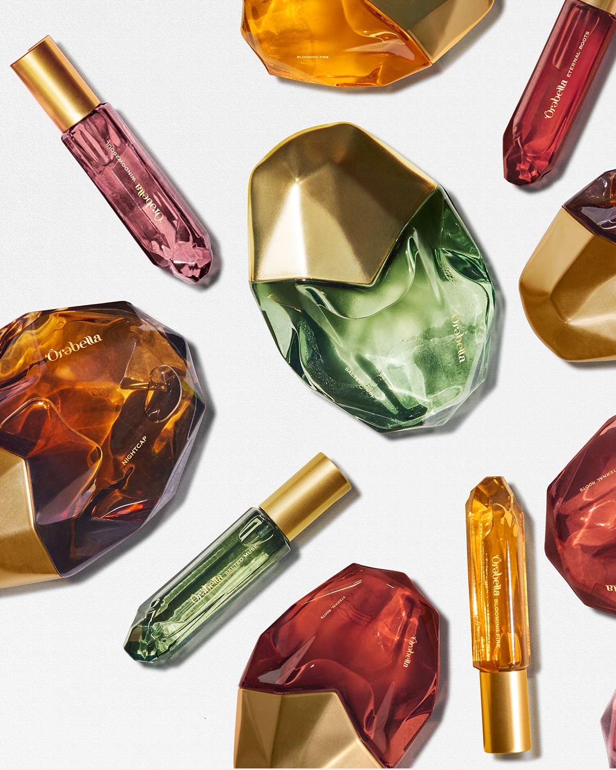 Įvairių spalvų „Örǝbella“ kvepalų buteliukai su auksiniais dangteliais išdėlioti ant balto paviršiaus, sukuria prabangos ir spalvų harmoniją.