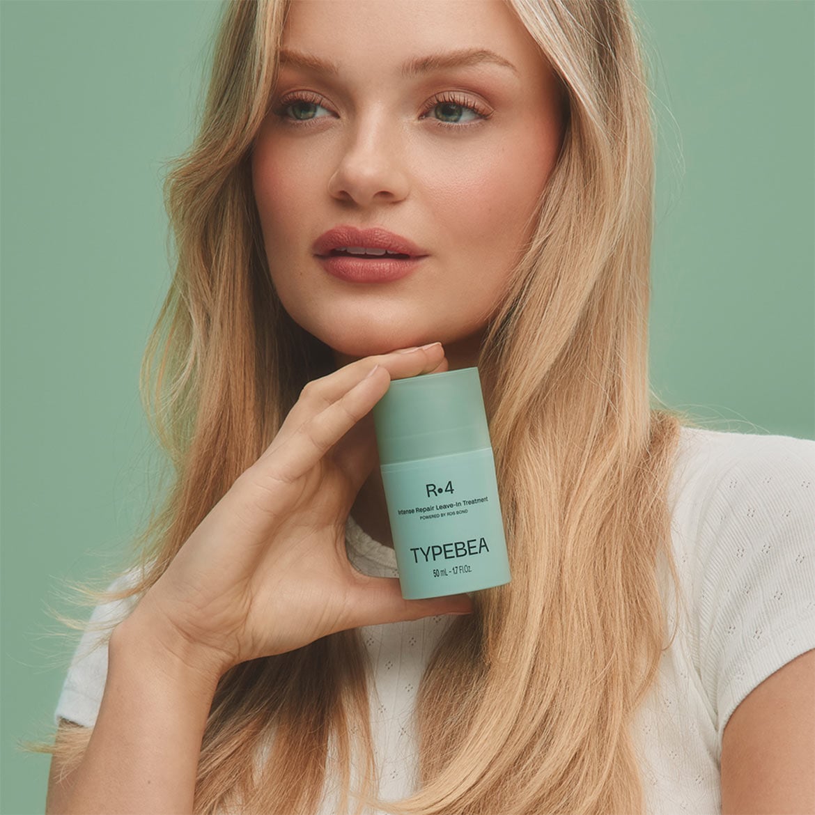 Šviesiaplaukė moteris laiko „Typebea R.4 Intense Repair Leave-In Treatment“ buteliuką, kurio spalva primena šviesią mėtų spalvą.