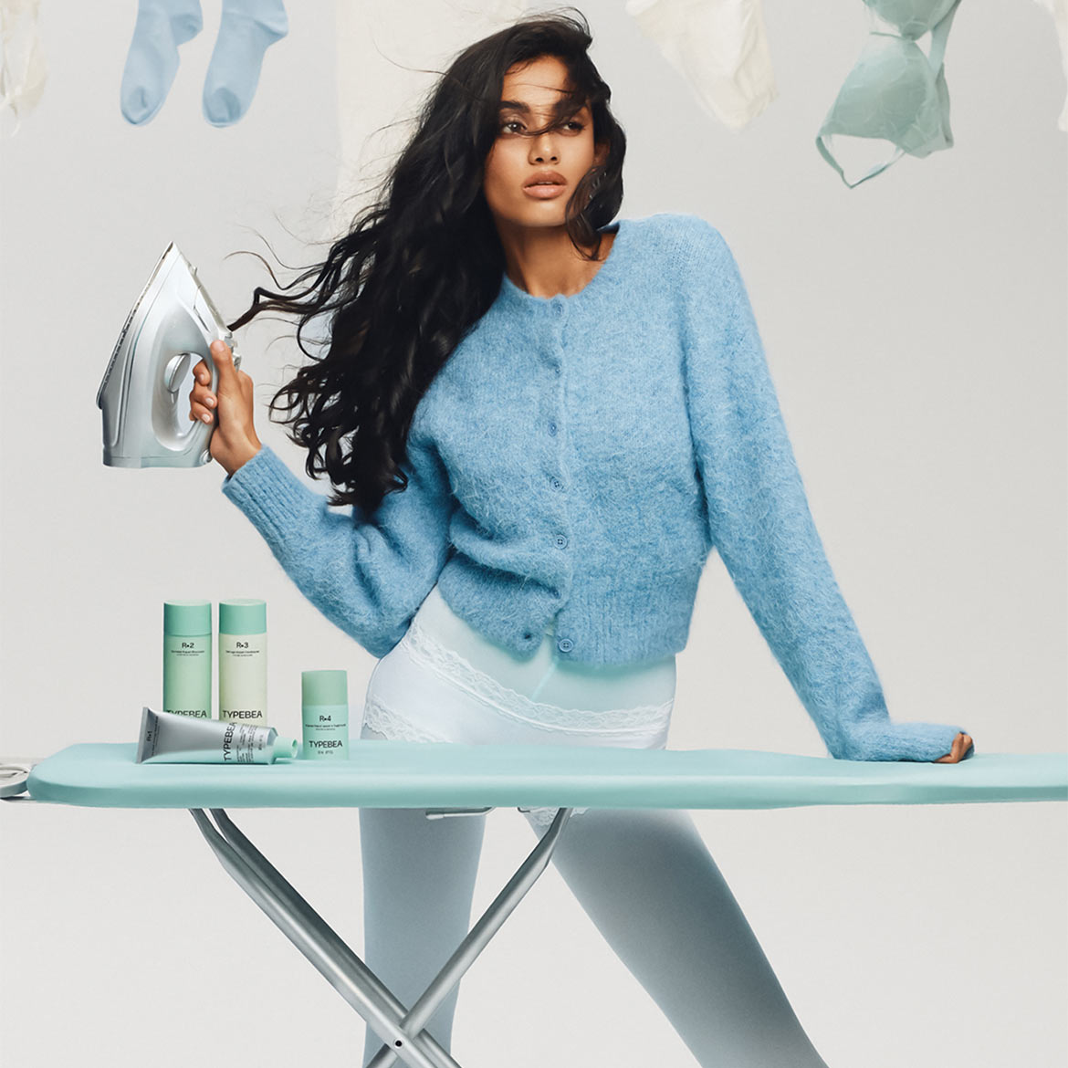 Nuotraukoje matome moterį su mėlynu megztiniu, laikančią lygintuvą, o ant lyginimo lentos išdėlioti „TYPEBEA“ odos priežiūros produktai.