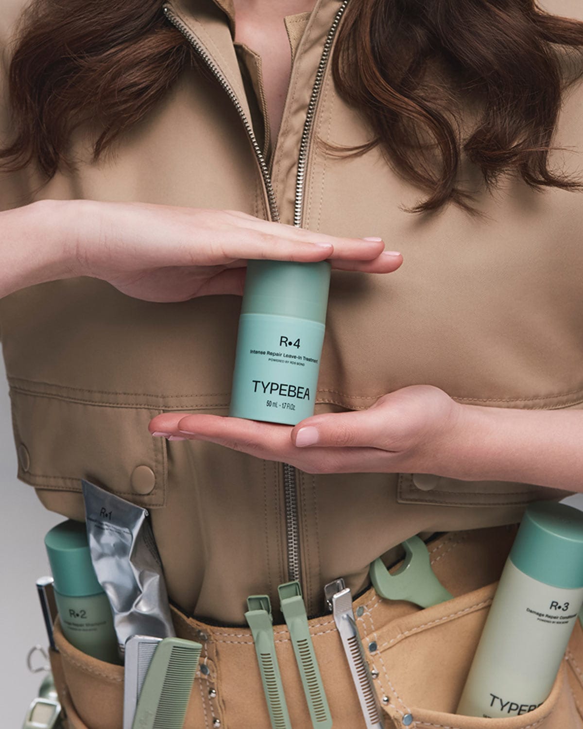 Moteris, vilkinti smėlio spalvos kombinezoną, laiko „Typebea R.4 Intense Repair Leave-In Treatment“ buteliuką, o jos įrankių dirže matyti kiti plaukų priežiūros produktai ir įrankiai.