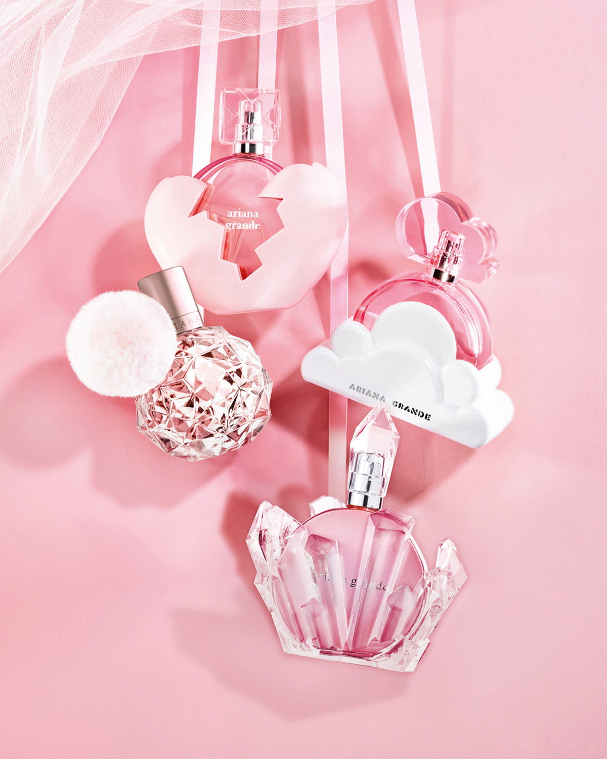 Keturi Ariana Grande kvepalų buteliukai su rožiniais atspalviais, pakabinti ant rožinių juostelių ir tiulio, sukuria žaismingą ir moterišką kompoziciją.
