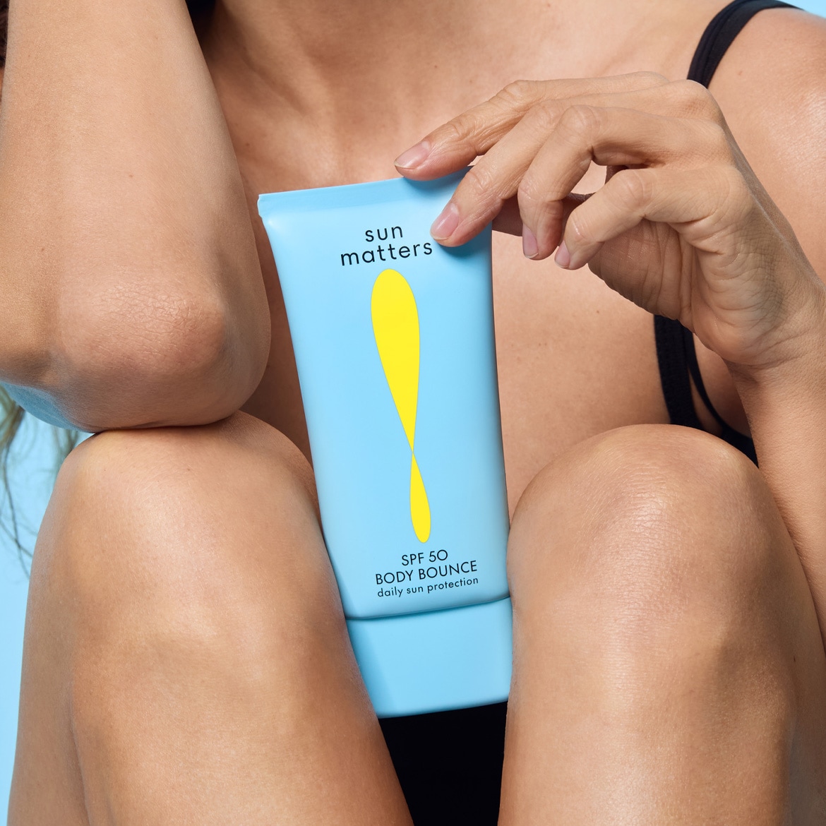 Žmogus rankoje laiko mėlyną Sun Matters SPF 50 Body Bounce kūno apsaugos nuo saulės kremo tūbelę stambiu planu prie odos.