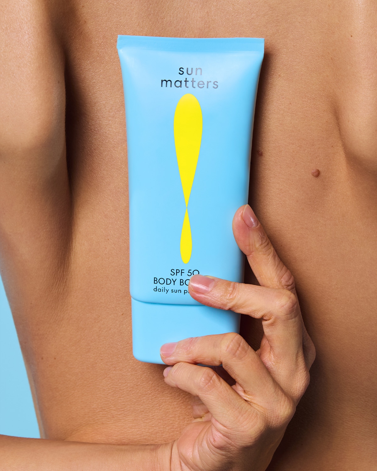Šviesiai mėlynas „Sun Matters“ SPF 50 kūno losjono buteliukas su geltonu lašo formos logotipu, kurį laiko ranka ant nuogos odos, užtikrina kasdienę apsaugą nuo saulės.