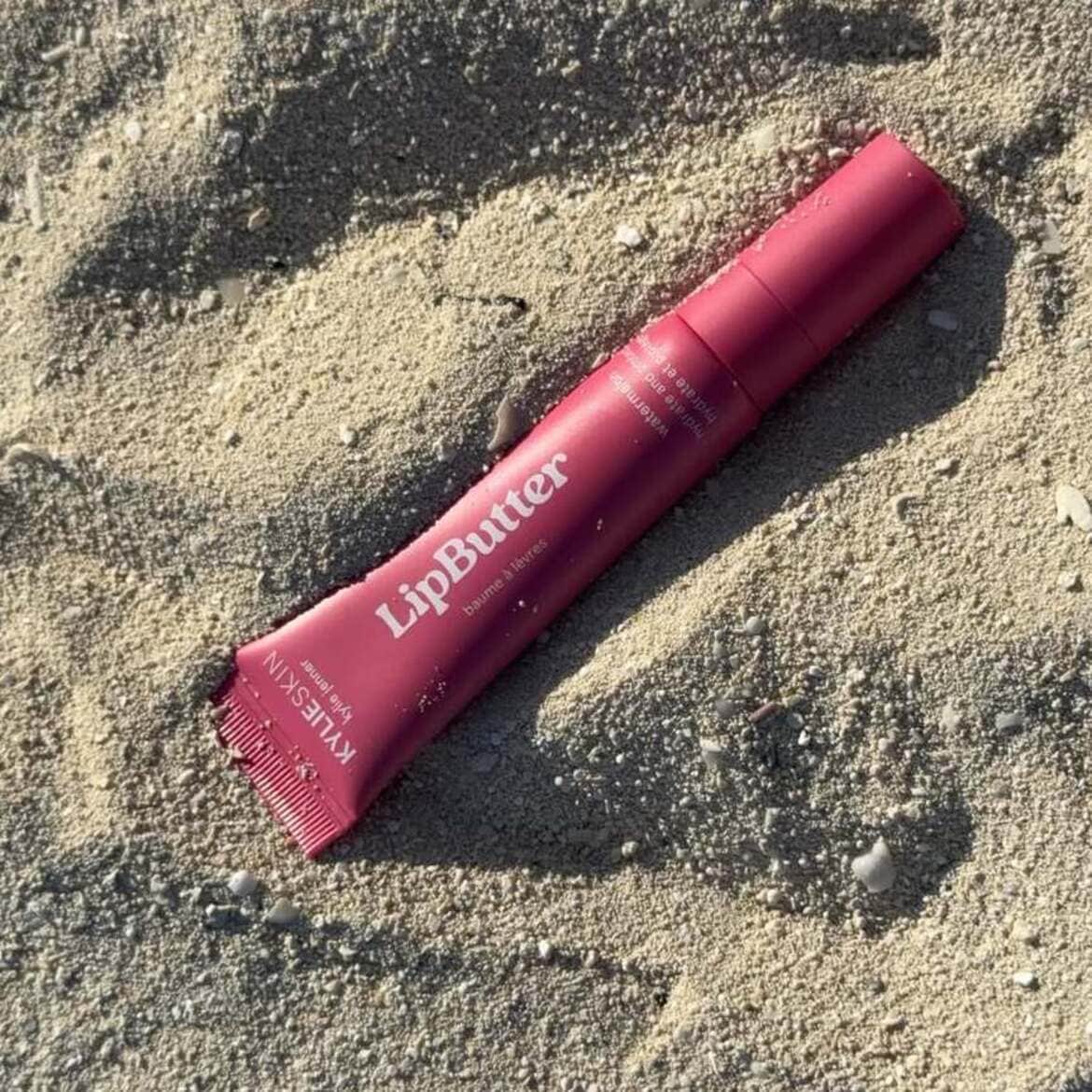 Kylie Skin lūpų sviestas, rožinės spalvos tūbelėje su baltu užrašu „LipButter“, guli smėlyje, o ant jo matyti smėlio kruopelės.