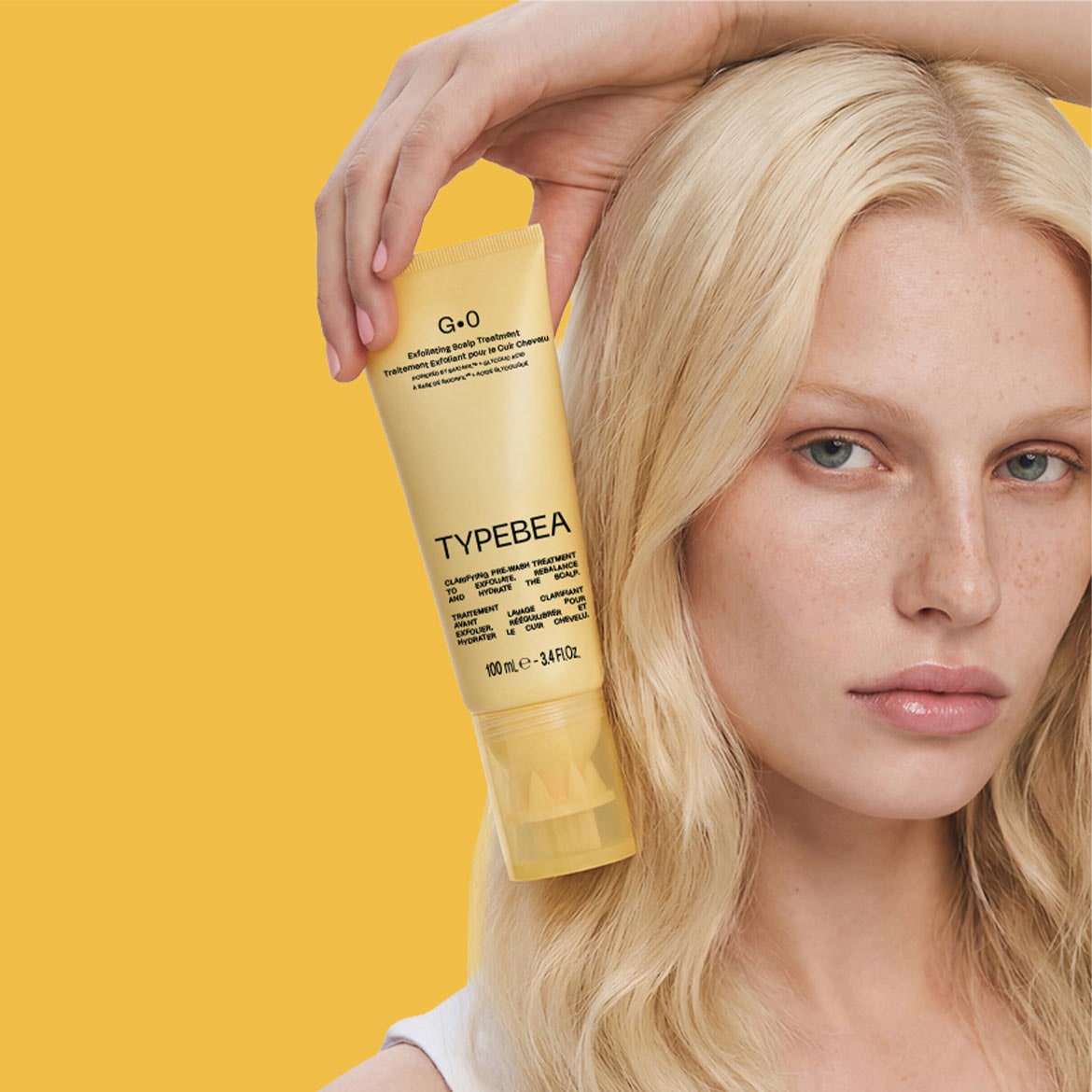 Šviesiaplaukė moteris laiko geltoną „TYPEBEA G.0 Exfoliating Scalp Treatment“ tūbelę, skirtą galvos odos šveitimui ir drėkinimui, kuri padeda atkurti pusiausvyrą.