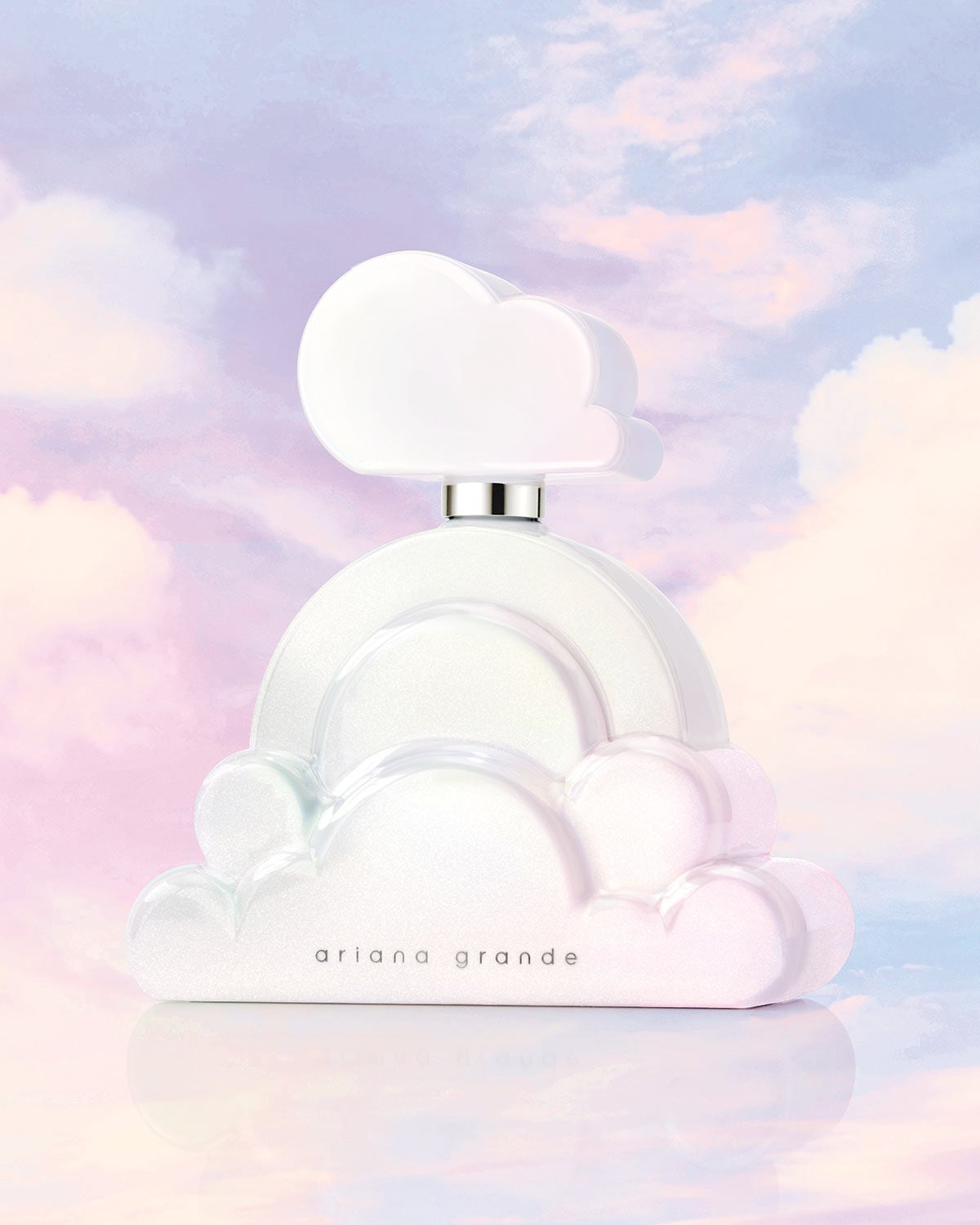 Ariana Grande Cloud kvepalai, baltas, debesies formos buteliukas su sidabriniu kakleliu ir užrašu „ariana grande“ ant buteliuko, pastatytas ant švelniai rožinio ir melsvo debesuoto fono.