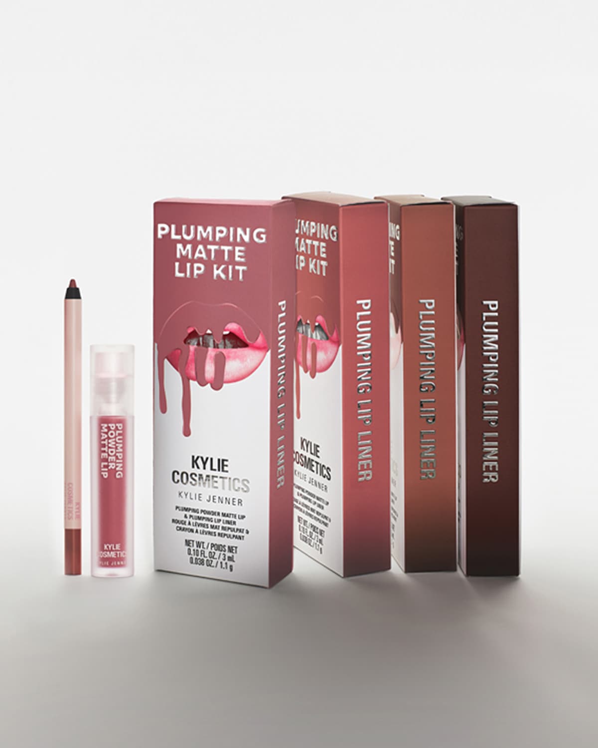 Kylie Cosmetics Plumping Matte lūpų rinkinys, kurį sudaro lūpų pieštukas ir skysti matiniai lūpų dažai, yra rožinės spalvos pakuotėje su lūpų atvaizdu, iš kurio laša lūpų dažai, o šalia stovi keturios rudos spalvos lūpų pieštukų dėžutės.