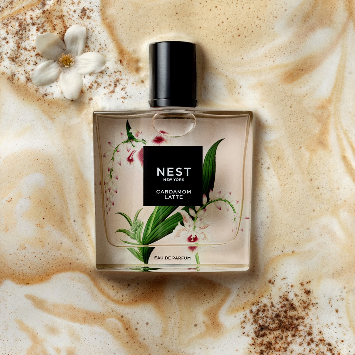 „Nest New York Cardamom Latte Eau de Parfum“ kvepalai, kurių buteliukas dekoruotas gėlių ir lapų motyvais, yra pastatytas ant kreminės kavos putos su cinamono pabarstukais, o šalia guli baltas žiedas.