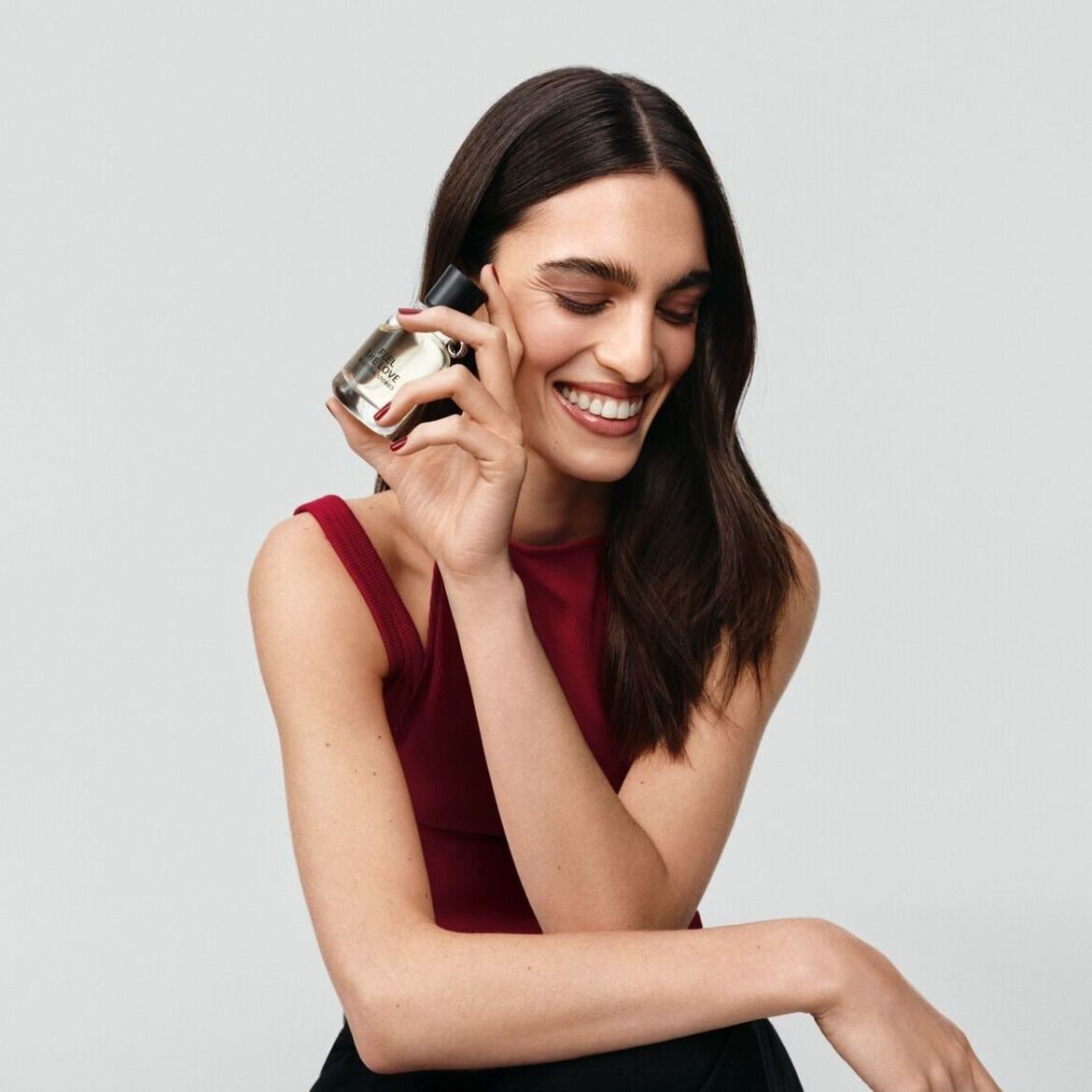 Švytinti moteris raudona palaidine laiko skaidrų kvepalų buteliuką su juodu dangteliu, užrašytu „First The Love Sutimers“ ir plačiai šypsosi.