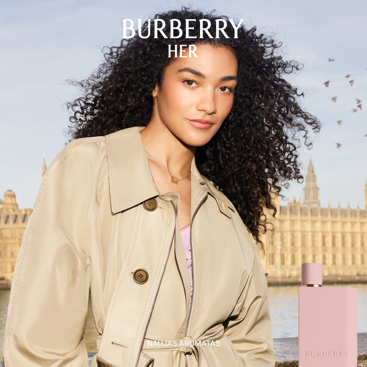 Burberry Her naujas aromatas: moteris su smulkiais garbanotais plaukais, vilkinti smėlio spalvos paltą, fone matosi Parlamento rūmai ir šviesiai rožinis Burberry kvepalų buteliukas.