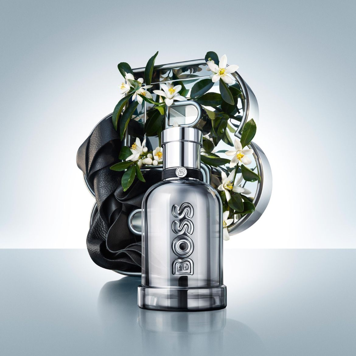 Sidabrinis „BOSS The Scent Magnetic“ kvepalų buteliukas su juodu odiniu dirželiu ir baltomis gėlėmis, atspindintis eleganciją ir magnetinę trauką.