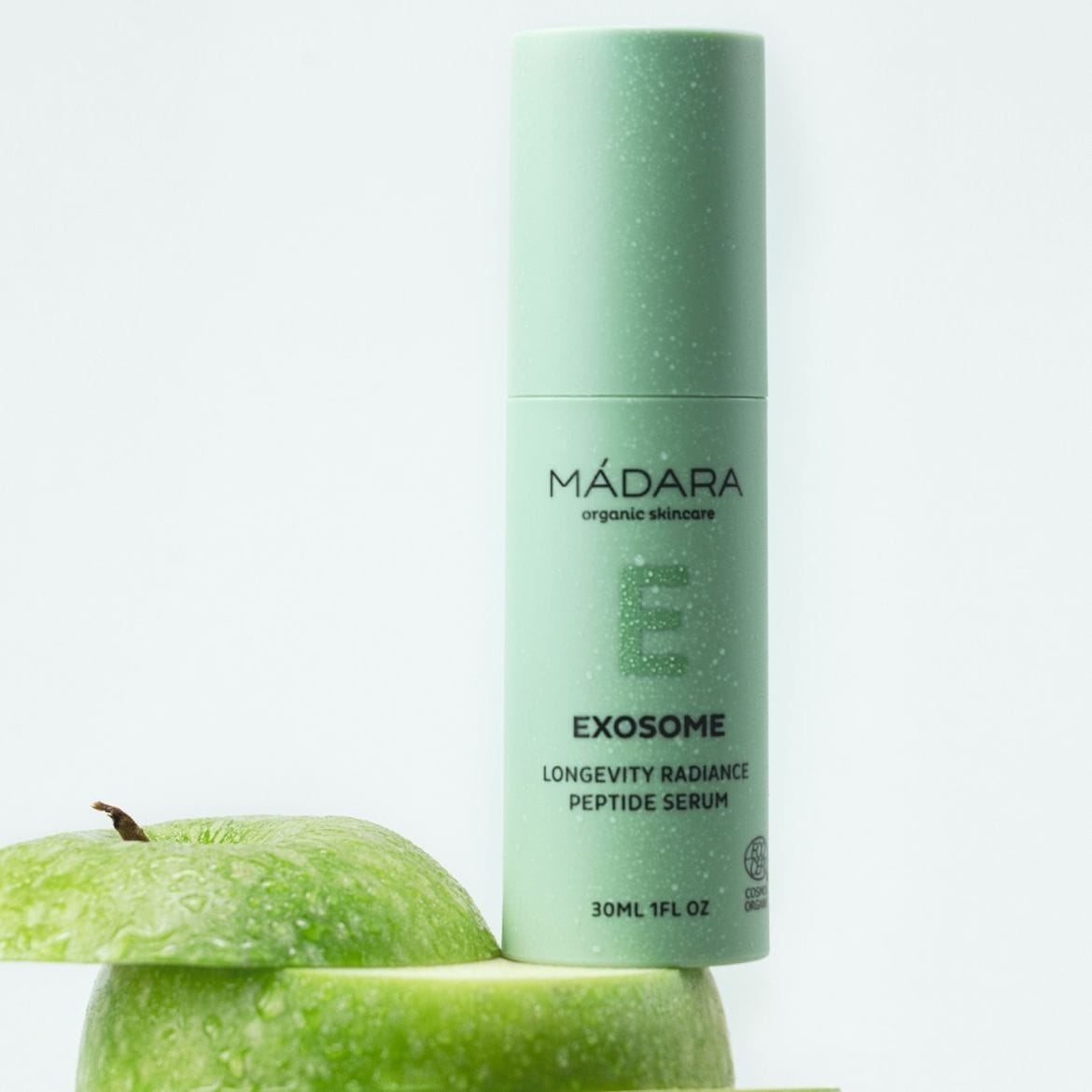 Šviesiai žalios spalvos „Mádara“ ekologiškos odos priežiūros priemonės „Exosome Longevity Radiance Peptide Serum“ buteliukas, kurio talpa 30 ml, stovi ant dviejų žalių obuolių griežinėlių.