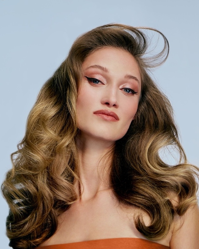 Moteris ilgais, banguotais plaukais stovi grakščiai, demonstruodama savo garbanotas garbanas ir pasitikėjimą savimi.