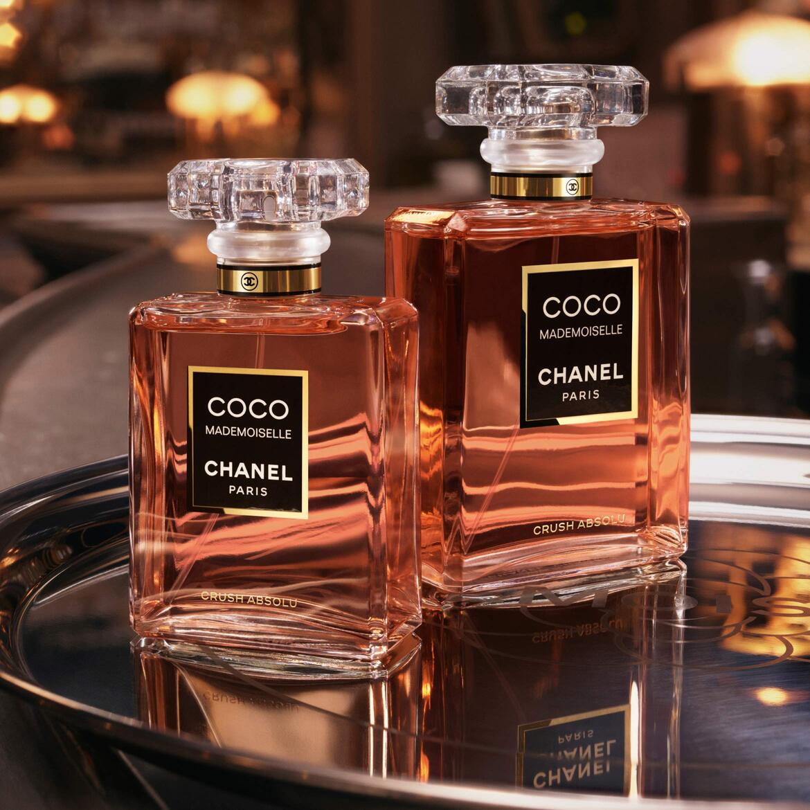 Du "Coco Mademoiselle Chanel Paris Crush Absolu" kvepalų buteliukai su skaidriais, raižytais kamšteliais ir auksiniais akcentais, užpildyti persikų spalvos skysčiu, stovi ant blizgančio sidabrinio padėklo, atspindinčio jų eleganciją.
