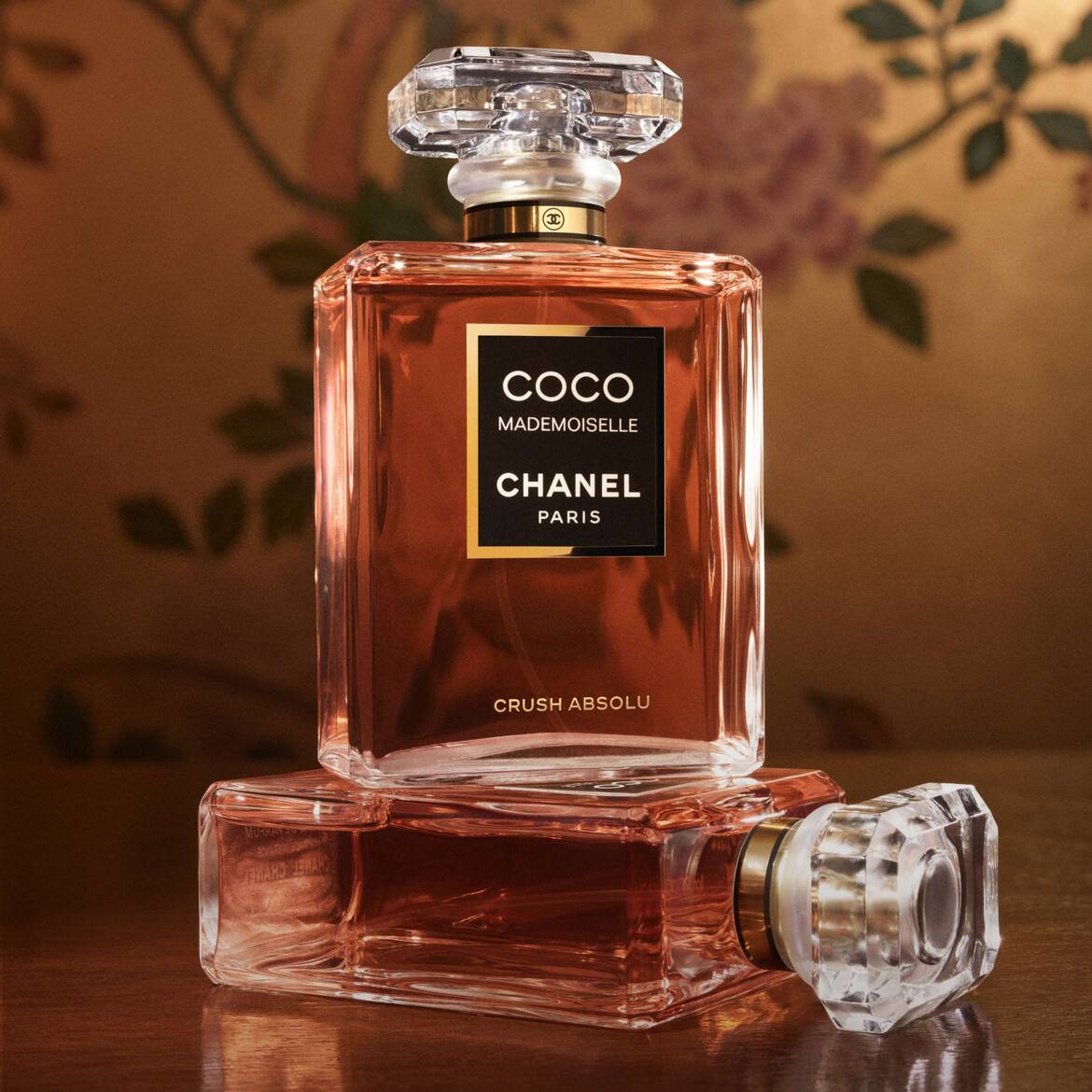„Coco Mademoiselle Chanel Crush Absolu“ kvepalų buteliukas su rausvu skysčiu ir skaidriu kristaliniu dangteliu, pastatytas ant kito, gulinčio buteliuko, sukuria elegantišką kompoziciją.