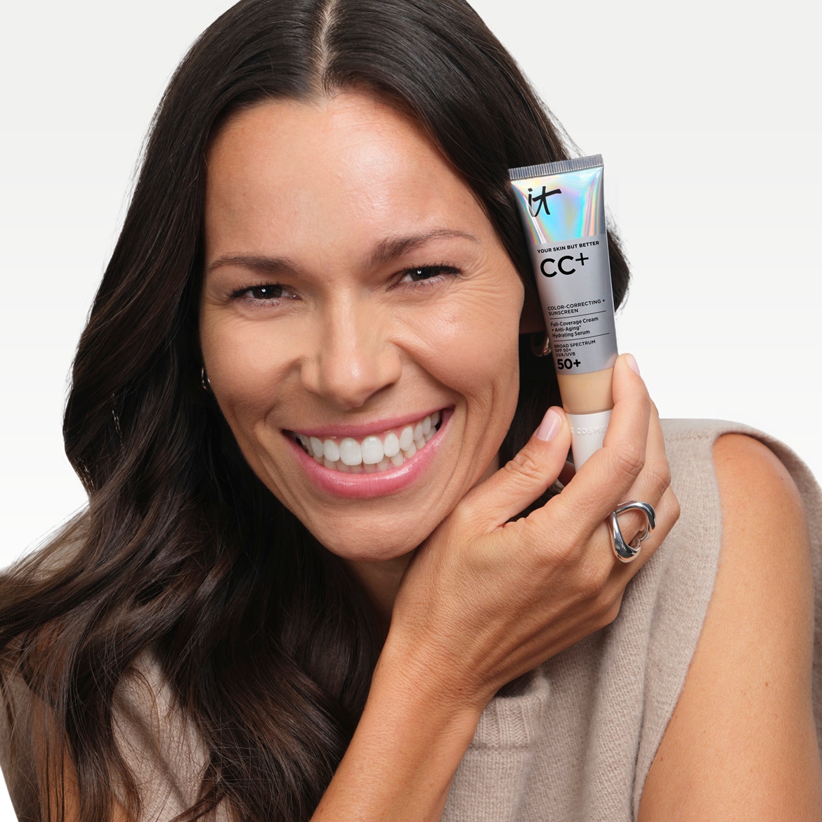 Moteris šypsosi ir laiko „IT Cosmetics CC+ Cream“ tūbelę, kuri yra odos spalvos, su užrašu „YOUR SKIN BUT BETTER CC+ COLOR-CORRECTING + SUNSCREEN SPF 50+“.