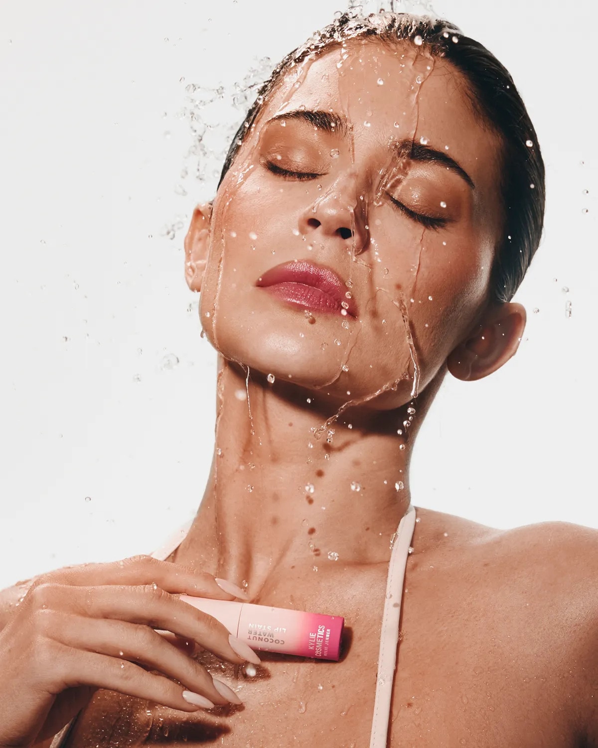 Asmuo su užmerktomis akimis stovi po vandens srove, laiko lūpų priemonę prie kaklo. Ant produkto matomas užrašas „Kylie Cosmetics Coconut Lip Stain“.