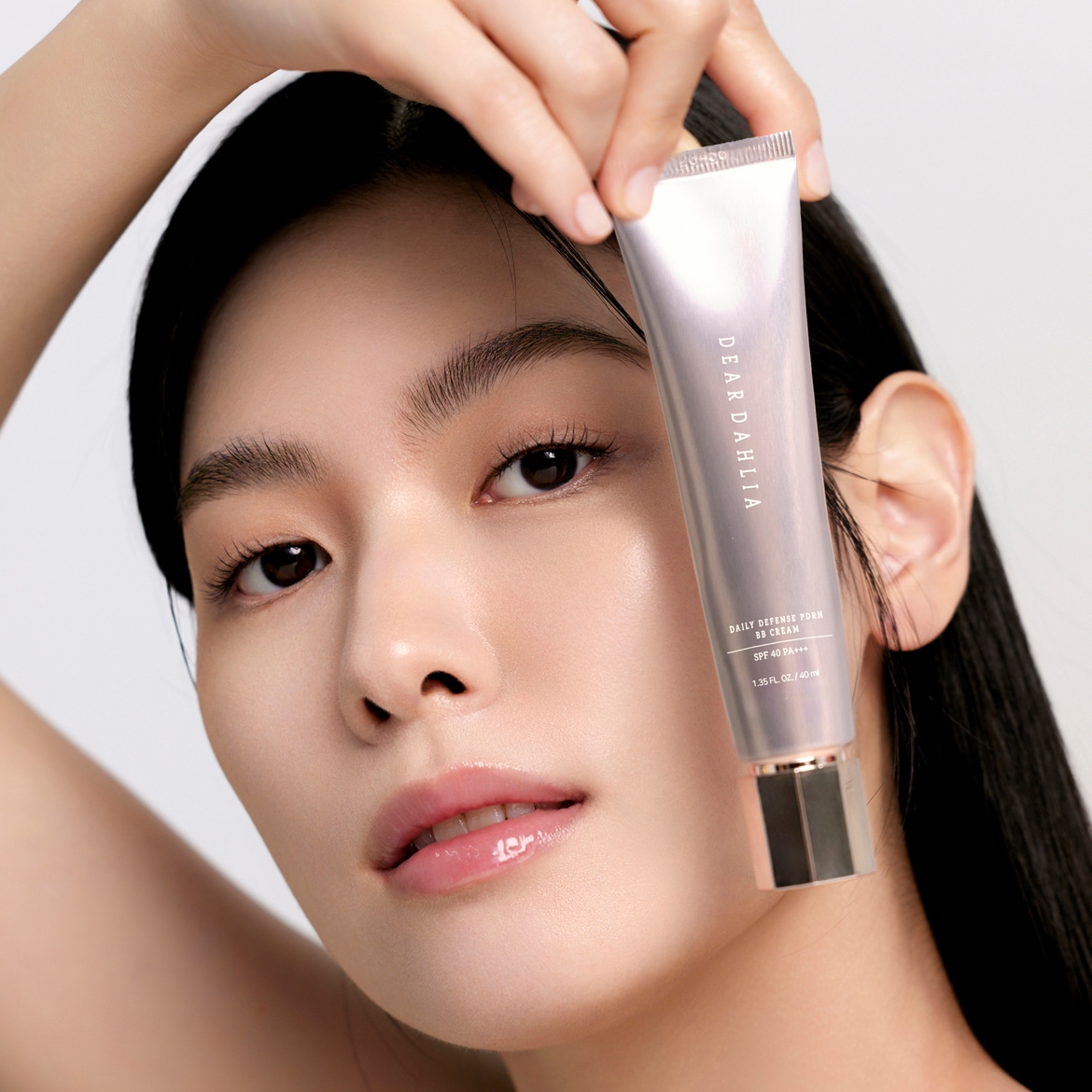 Moteris laiko „Dear Dahlia Daily Defense PDRN BB Cream SPF 40 PA+++“ tūbelę, kuri yra šviesiai violetinės spalvos su metaliniu dangteliu, o jos veidas atrodo spindintis ir natūralus.