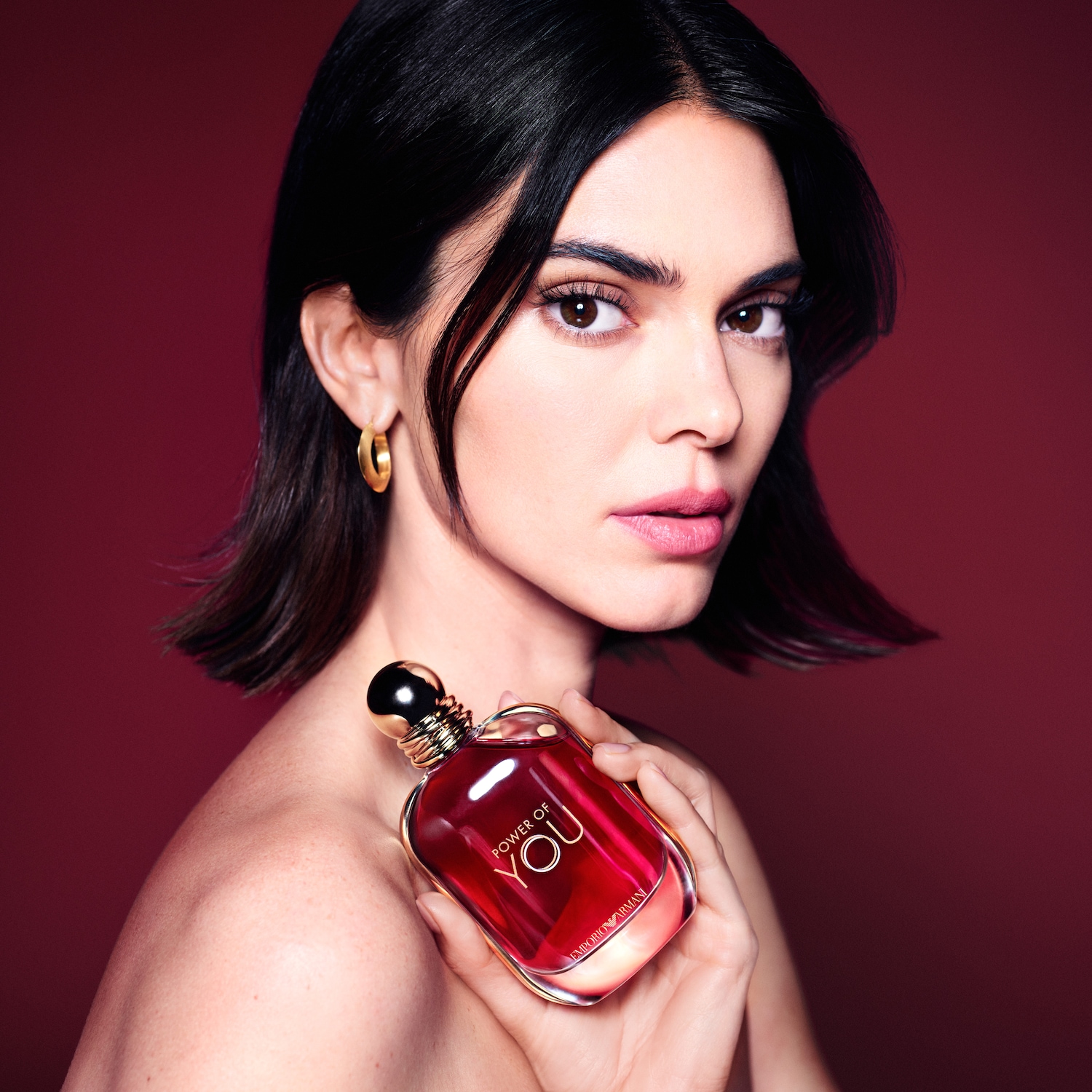 Šviesi moteris su trumpais tamsiais plaukais laiko raudoną „Emporio Armani Power of You“ kvepalų buteliuką su auksiniu dangteliu ant raudono fono.