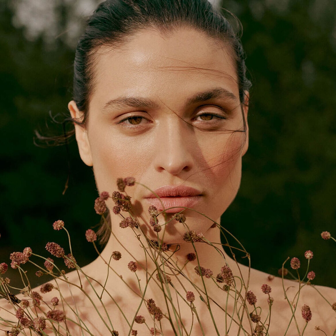 Portretas moters su šviesia oda, rudomis akimis ir plaukais, surištais į kuodą, veidą dengia smulkūs lauko augalai.