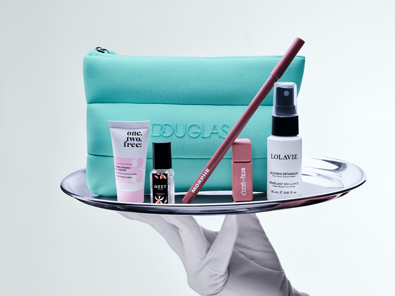 A silver tray held by a gloved hand showcases a turquoise Douglas pouch, One.Two.Free! face cream, Nest perfume, Morphe lip liner, About-Face lip product, and Lolavie detangler.
