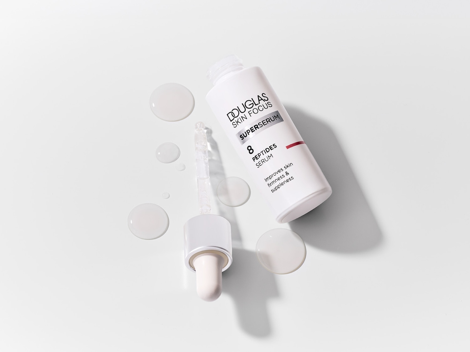 „Douglas Skin Focus Superserum 8 Peptides Serum“ baltas buteliukas su pipete ir išpiltu serumu, kuris pagerina odos stangrumą ir elastingumą.