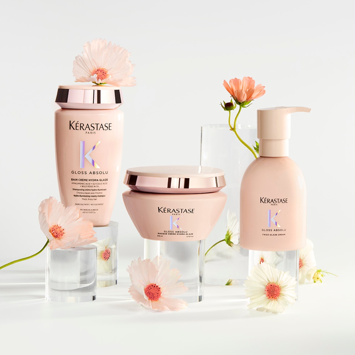 Šampūnas, kaukė ir kremas Kérastase Gloss Absolu serijos, šviesiai rožiniuose buteliukuose, skirti drėkinti ir suteikti plaukams spindesio, papuošti gėlėmis.