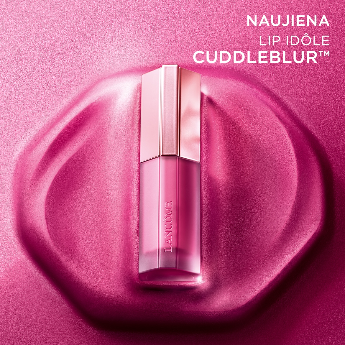 Naujiena! Rožinis „Lancôme Lip Idôle Cuddleblur™“ lūpų blizgesys su matiniu dangteliu ir skaidriu korpusu, patalpintas rausvame fone.