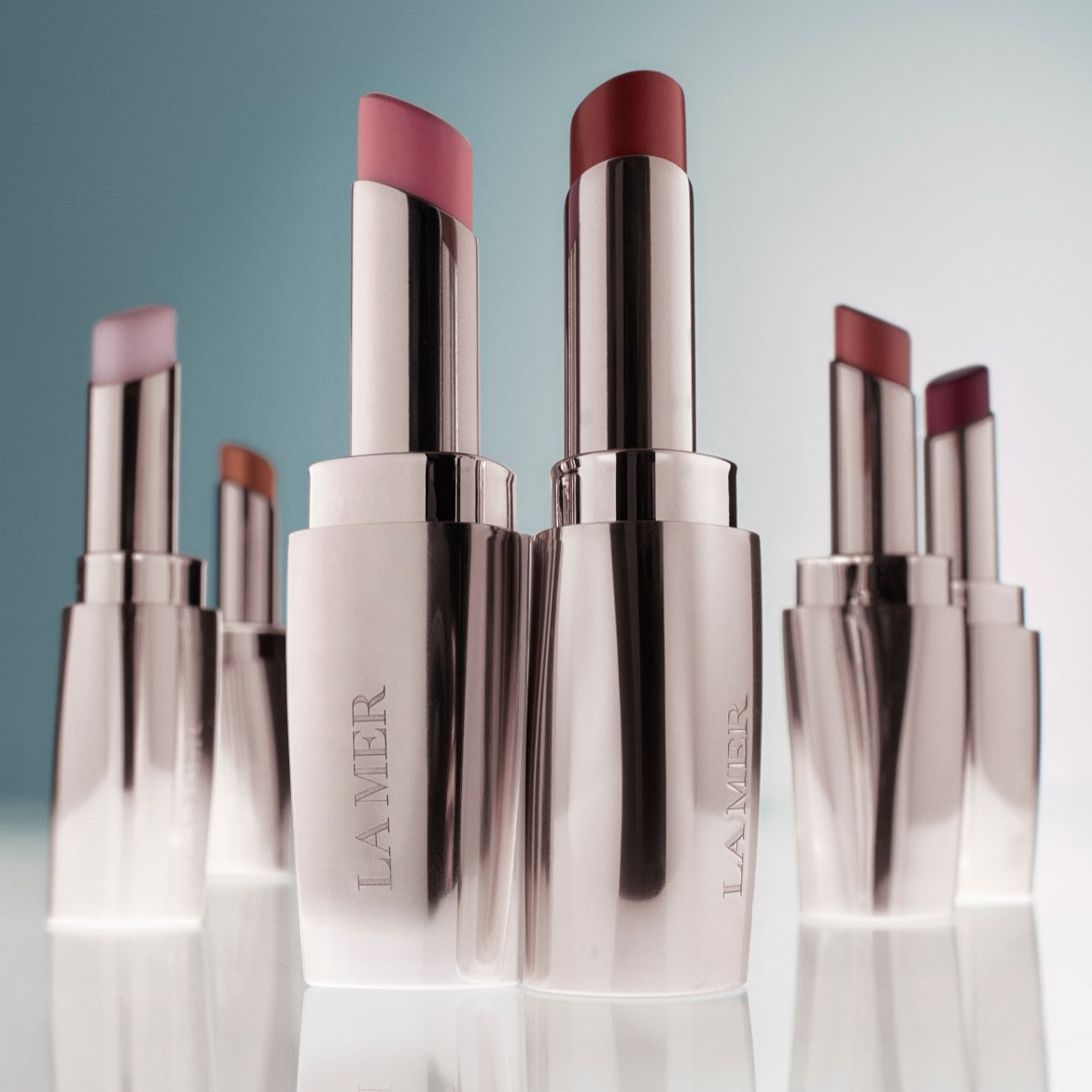 Štai „La Mer“ lūpų balzamai sidabrinėje pakuotėje su atsuktais įvairių atspalvių lūpų dažais, nuo šviesiai rausvos iki tamsiai raudonos.