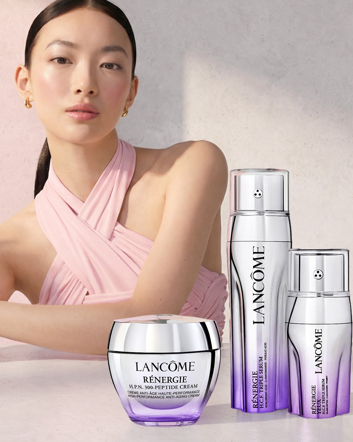 Elegantiška moteris su švelniai rausva suknele ir auksiniais auskarais, švelniai žvelgianti į priekį, o šalia jos – „Lancôme Rénergie H.P.N. 300-Peptide Cream“ ir „Rénergie H.C.F. Triple Serum“ produktai.