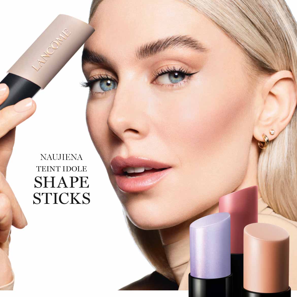 Šalia moters veido matomi „Lancôme Teint Idole Shape Sticks“ makiažo pagrindai, kurių atspalviai yra švelniai violetiniai, rožiniai ir smėlio spalvos.