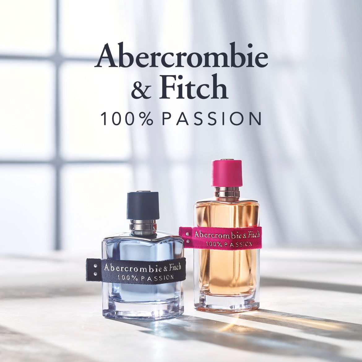 „Abercrombie & Fitch 100% Passion“ moteriški ir vyriški kvepalai ant šviesaus fono, šalia didelio prekės ženklo pavadinimo.