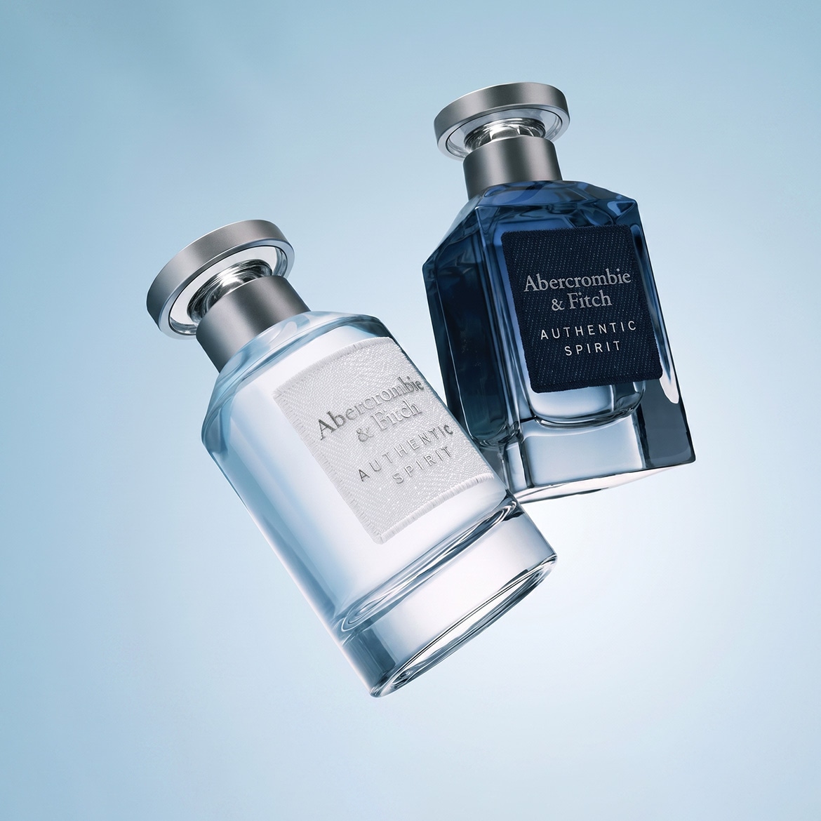Du Abercrombie & Fitch Authentic Spirit kvepalų buteliukai, vienas skaidrus su baltu užrašu, kitas tamsiai mėlynas su juodu užrašu, plūduriuoja šviesiai mėlyname fone.