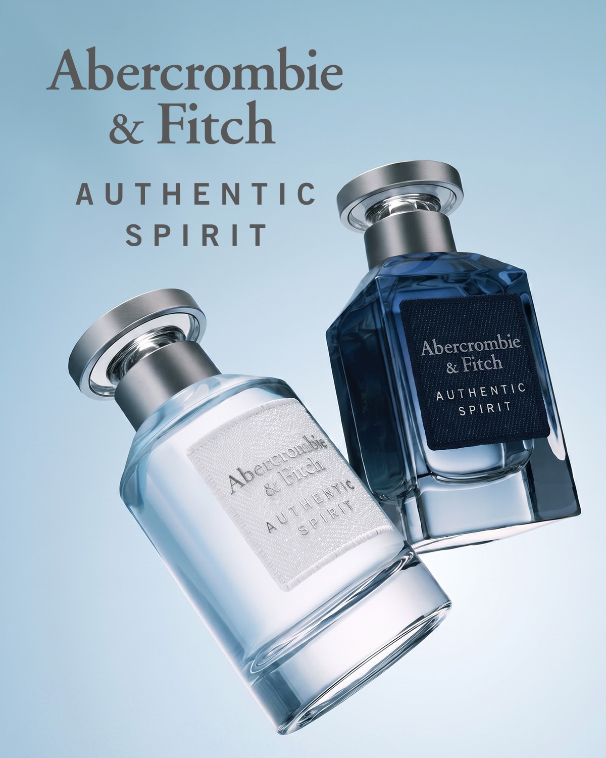 Du Abercrombie & Fitch Authentic Spirit kvepalų buteliukai, vienas skaidrus su baltu užrašu, kitas tamsiai mėlynas su juodu užrašu, abu su sidabriniais dangteliais, elegantiškai pakreipti šviesiai mėlyname fone.