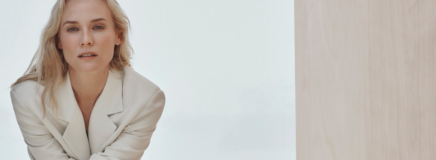 Šviesiaplaukė moteris, vilkinti kreminės spalvos švarką, žiūri tiesiai į priekį ramiu ir pasitikinčiu žvilgsniu.