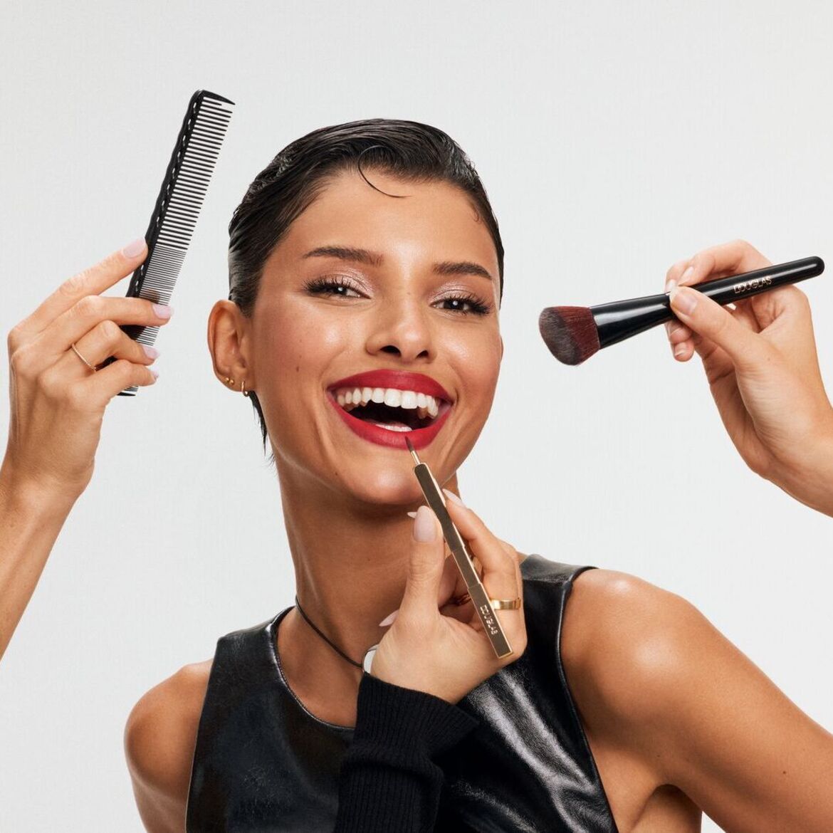 Švytinti moteris su raudonu lūpdažiu šypsosi, kol stilistai šukuoja jos plaukus šukomis ir tepa lūpas auksiniu lūpų pieštuku bei skruostus skaistalų teptuku.