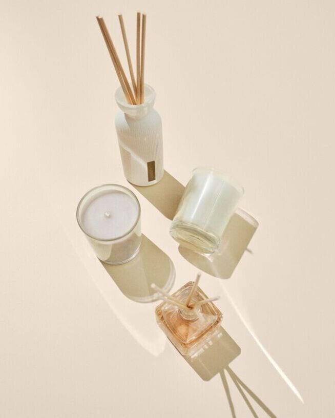 Nuotraukoje matomi kvapų difuzoriai su lazdelėmis ir žvakės stikliniuose induose, išdėstyti ant šviesaus fono.
